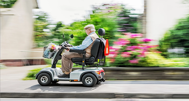 Elderly man in a cap driving a mobility scooter on a sidewalk with flowers in the background.