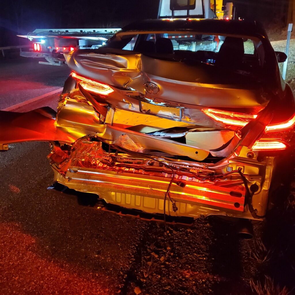 A rear view of a heavily damaged car with its back crushed and lights on. The vehicle is on the side of the road at night, with a tow truck visible in the background.