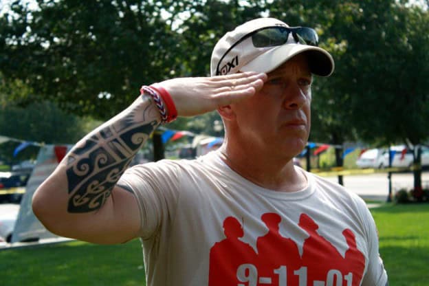 A man wearing a beige cap and sunglasses salutes outdoors. He has a tattoo on his left arm and is dressed in a shirt commemorating "9-11-01." Trees and a parked car are visible in the background.