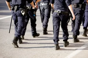 A group of police officers in uniform walk on an asphalt road, with only their lower bodies visible. All officers are wearing dark pants, belts with handcuffs, and other equipment.