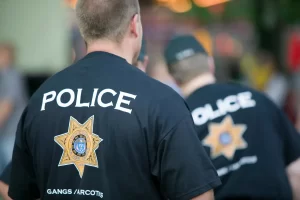 Two police officers are seen from behind, each wearing a black shirt labeled "POLICE" with an emblem and "Gangs/Narcotics" text on the back.