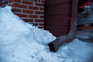 A brown downspout directs water into a pile of snow next to a brick wall, with part of a car visible in the background.