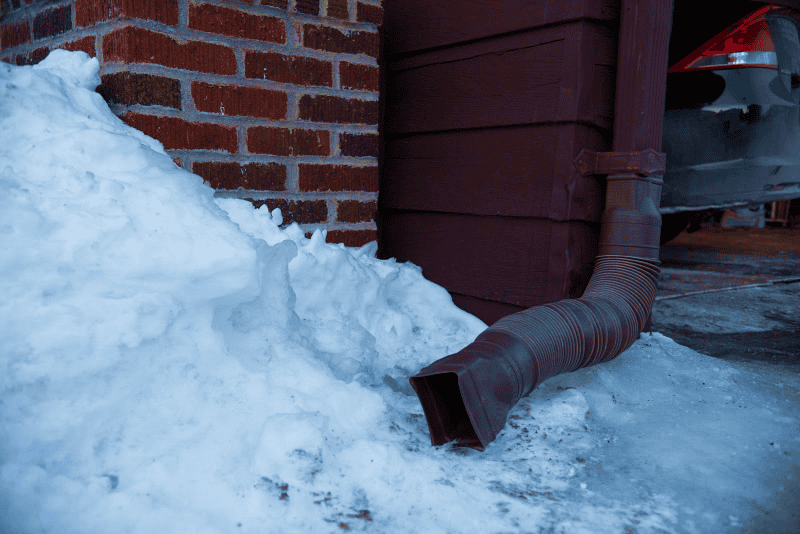 A brick wall with a brown downspout draining melted snow onto an icy patch by the side of a red-brown wooden structure. Snow is piled up around the base. A car is partially visible on the right.