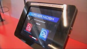Touchscreen waiver kiosk with a prompt to register. The screen displays icons of safety helmets for existing and new accounts, with instructions to input each participant individually.