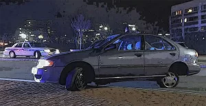 A sedan with a damaged front wheel is parked partially on the sidewalk at night as a police car with flashing lights is stationed nearby.