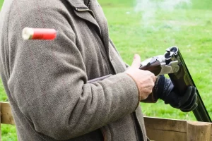 A person wearing a tweed jacket and black gloves ejects a spent shotgun shell from a double-barreled shotgun outdoors.
