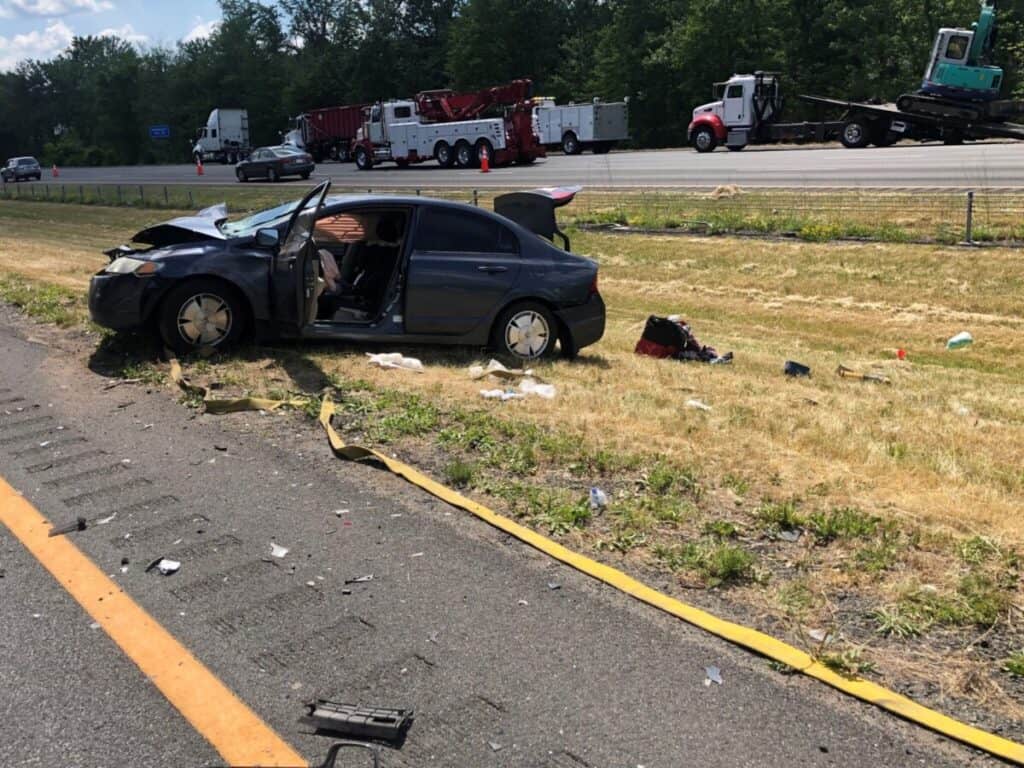 A damaged car is seen on the grass beside a highway with debris scattered around. Two tow trucks are visible in the background on the highway.