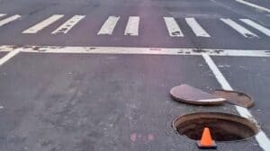 A street scene shows a crosswalk with a manhole cover off to the side and an orange traffic cone placed in front of the open manhole.