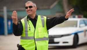 A police officer wearing a neon safety vest and sunglasses directs traffic with a smile, with a police car parked in the background.