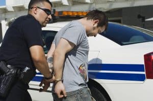 A police officer is handcuffing a man in front of a police car. The man has tattoos on his arm and is wearing a gray t-shirt.