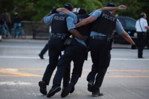 Three police officers in uniform assist each other while walking on a street. The central officer appears to be supported by the other two.