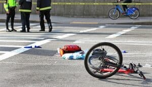 A bicycle lies overturned in a crosswalk, with scattered belongings nearby. Police officers are seen in the background near caution tape.