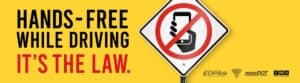 A road sign shows a hand holding a phone with a red no symbol. Text reads, "HANDS-FREE WHILE DRIVING IT'S THE LAW." Government logos are at the bottom right.