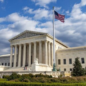 The United States Supreme Court building under a partly cloudy sky with the American flag flying in the foreground.