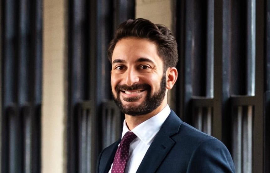 Man in a suit and tie smiling, standing in front of a dark, windowed background.