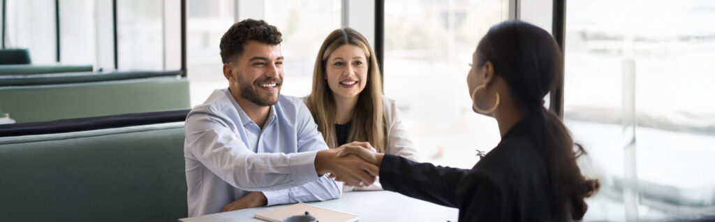 Three people in a business meeting. Two are seated, one shakes hands with a standing person across a table.