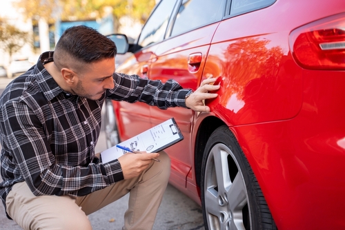 man crouching beside car inspecting vehicle damage while holding clipboard