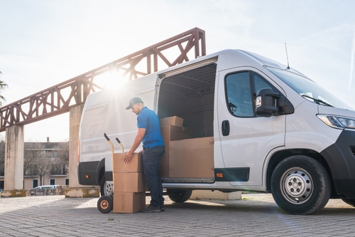 Delivery driver unloading stacked boxes from cargo van with hand truck