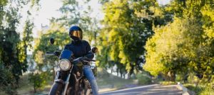 Motorcyclist wearing helmet, sunglasses, leather jacket, and jeans beside motorcycle