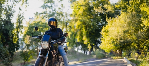 Motorcyclist wearing helmet, sunglasses, leather jacket, and jeans beside motorcycle