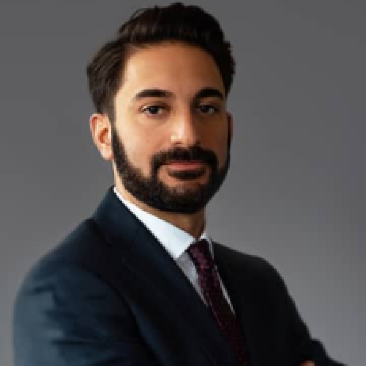 A man with dark hair and beard, in a suit and tie, stands against a plain background.
