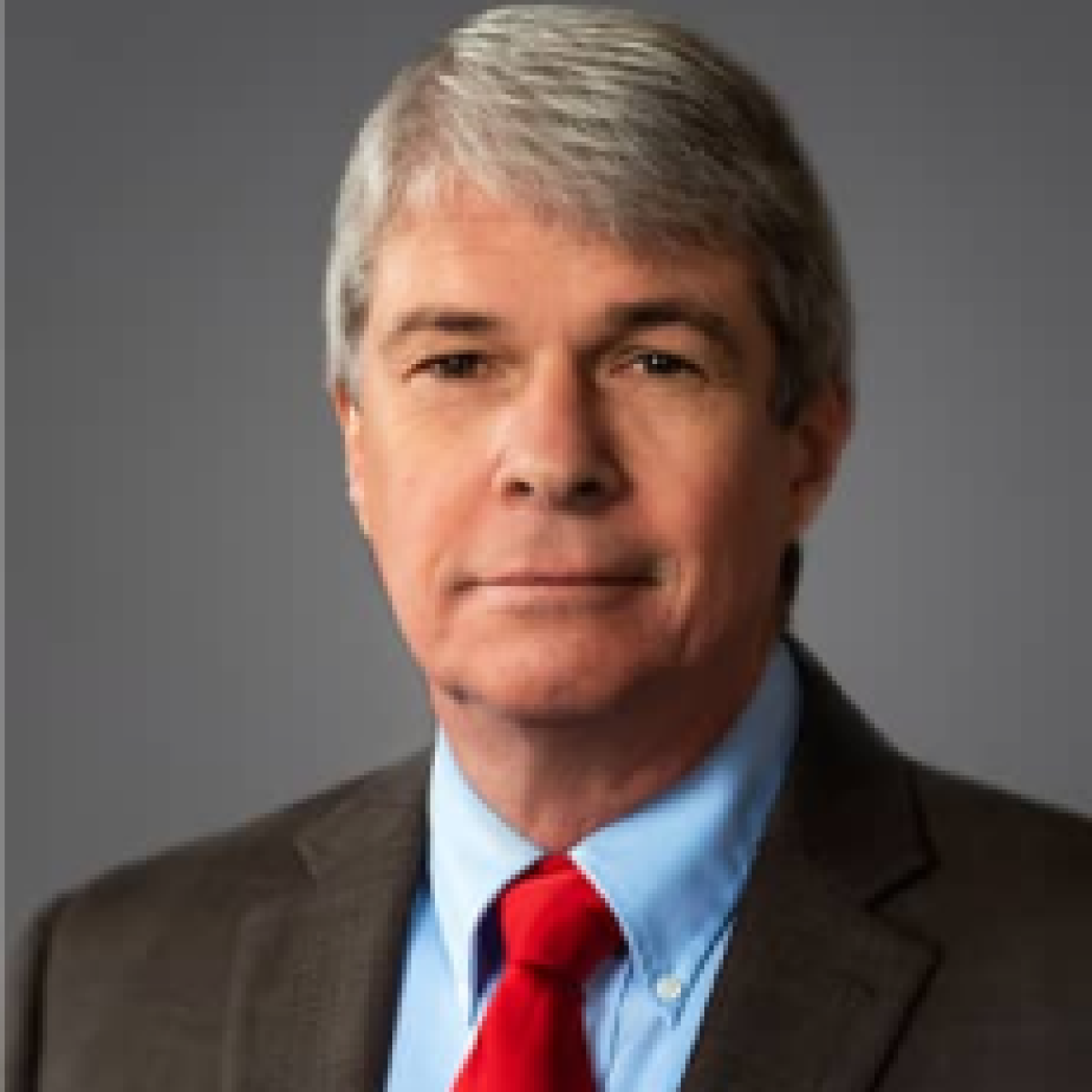 A man with gray hair wearing a brown suit, blue shirt, and red tie is posing against a gray background.