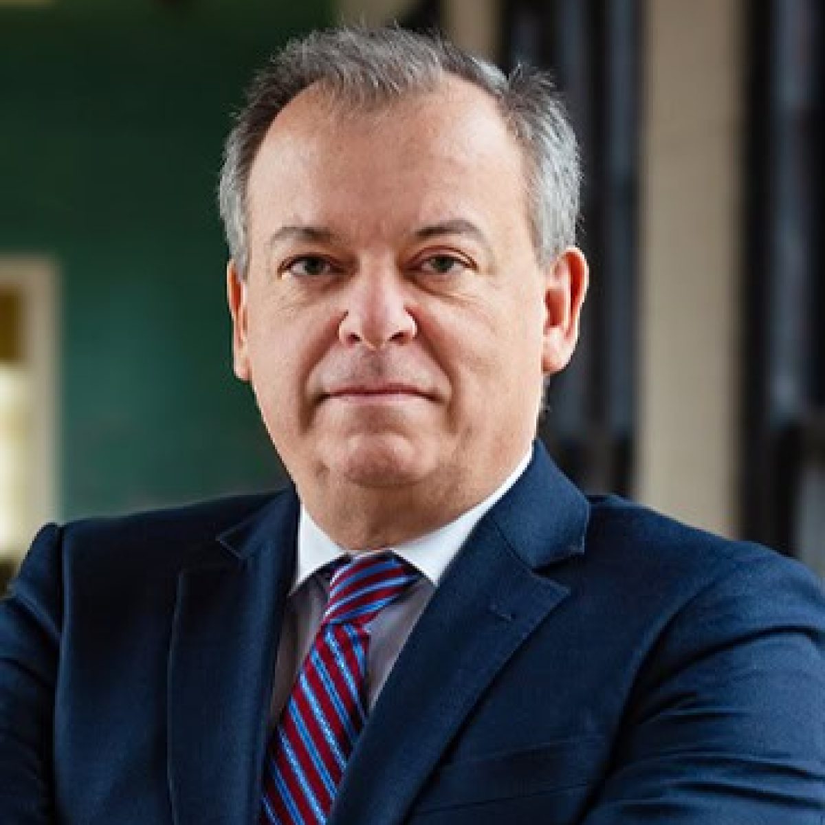 A middle-aged man with short gray hair wearing a dark suit, blue shirt, and red striped tie stands in front of a blurred indoor background.