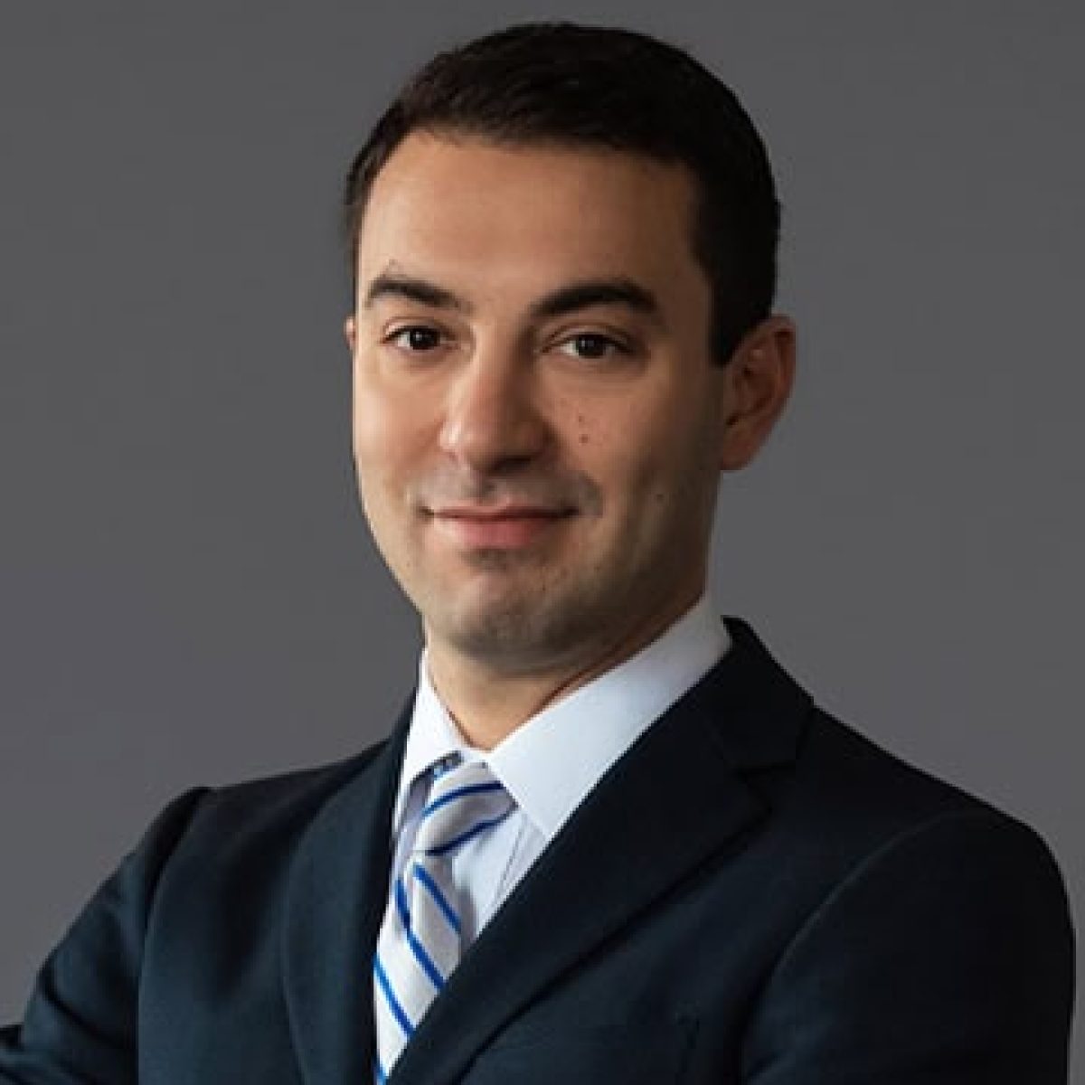 A man in a dark suit with a light blue striped tie poses confidently against a gray background.